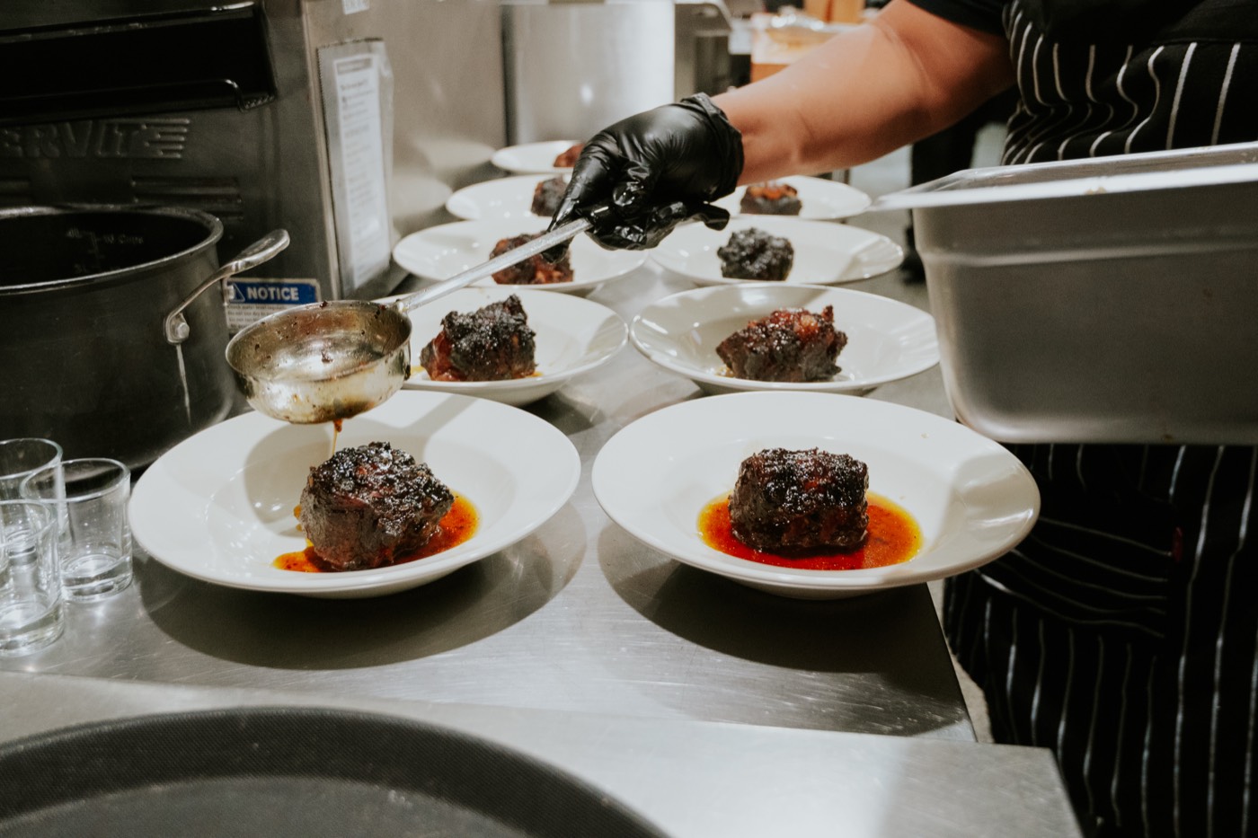 A gloved hand finishing plates of braised beef on the pass