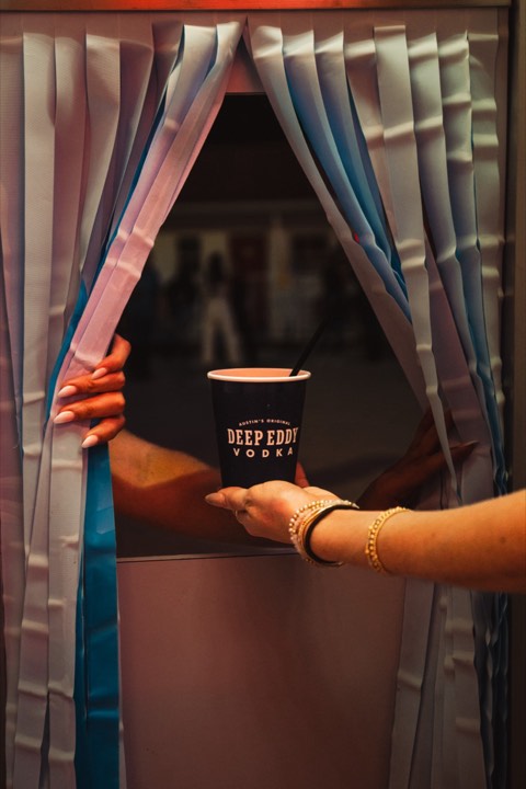 A hand passing a Deep Eddy cup through a curtained serving window